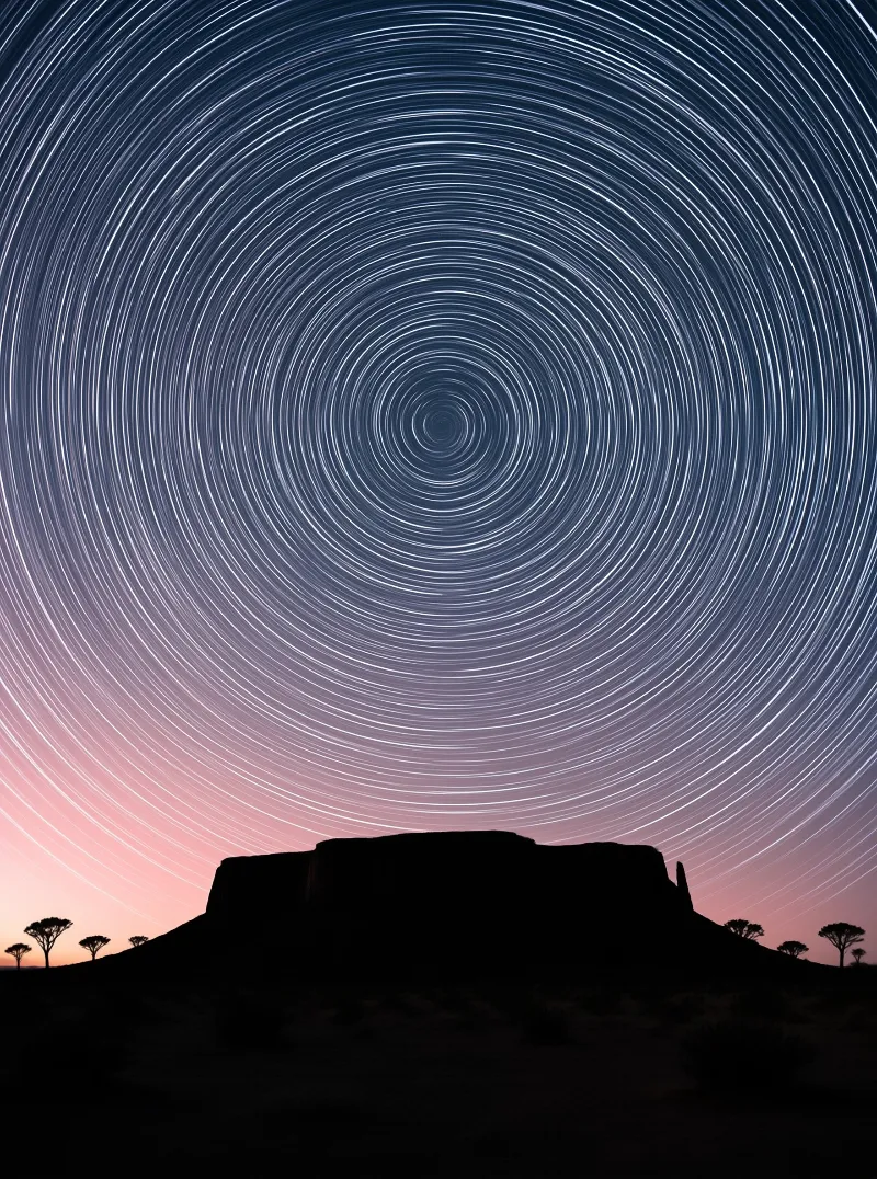 Spiraling star trails arc over a dark plateau dotted with umbrella trees under a pink horizon glow