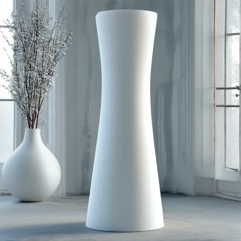 A tall white pedestal stands next to a vase with frosted branches in a bright minimalist room.