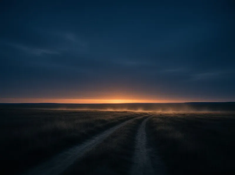 A worn dirt path curves across a dark grassland while low fog glows under the bright orange horizon.