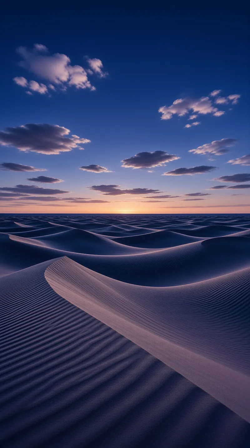 Wind-carved sand ridges roll toward a setting sun beneath a deep blue sky scattered with rose-tinted cloud.