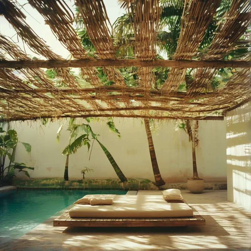 Wooden platform with pillows beneath thatched roof near turquoise pool in dappled sunlight.