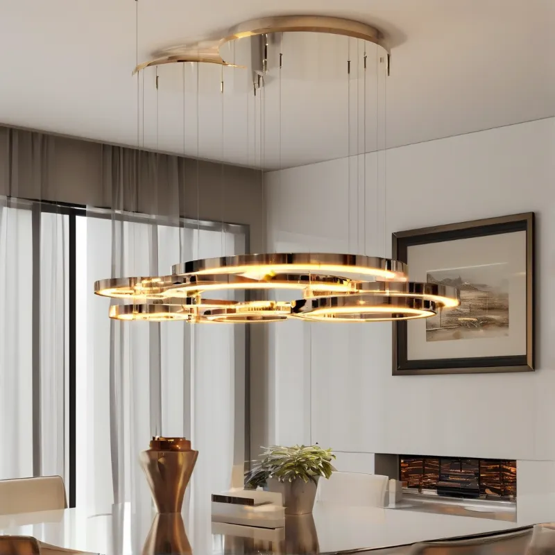 A tiered metallic ceiling fixture glows warmly above a reflective dining table within a minimalist white interior space.