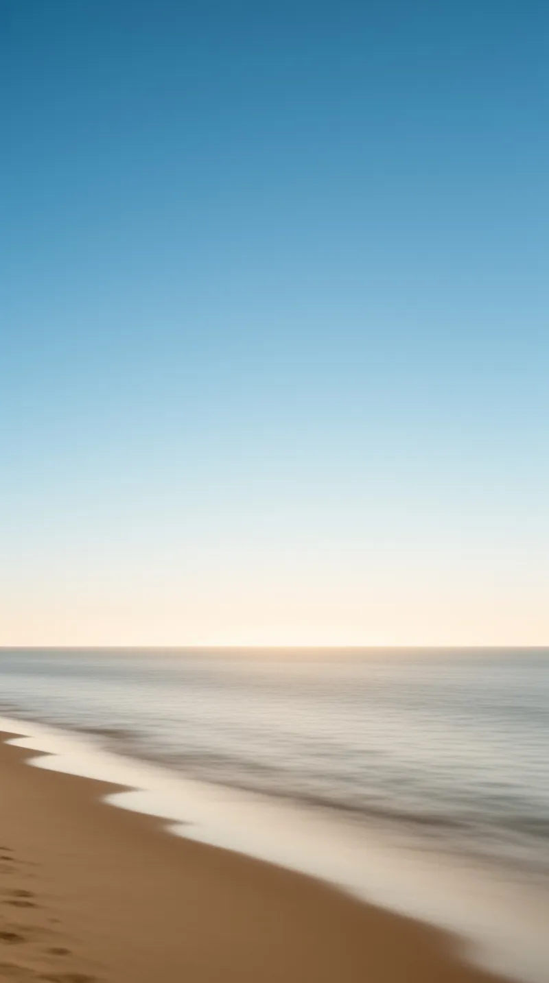 Gentle waves lap a smooth sandy shore beneath a clear sky that fades from blue to warm pale light.