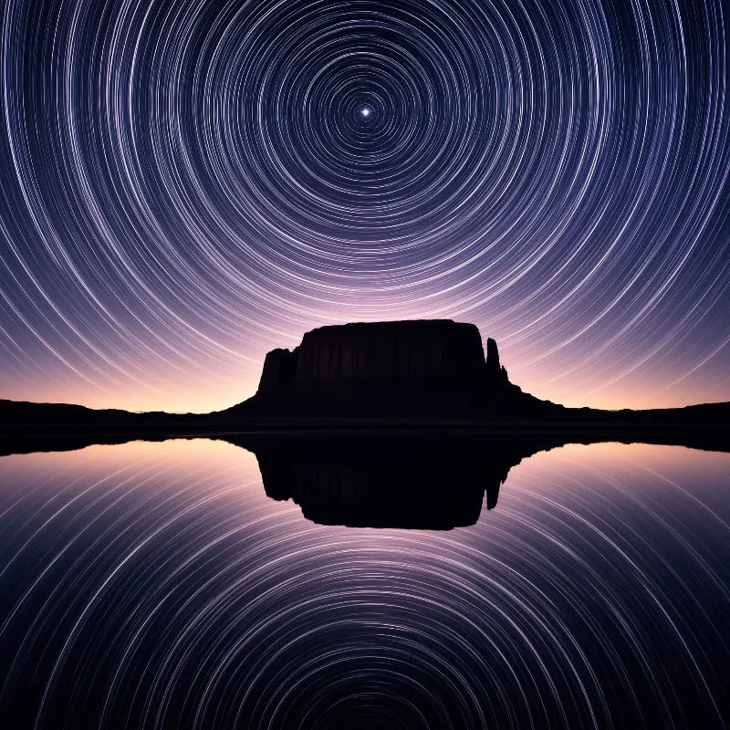 A dark mesa silhouette is centered beneath swirling concentric star trails that mirror perfectly in the calm water.