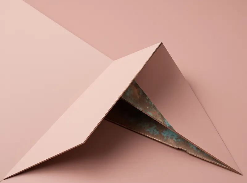 Folded copper sheets with blue green oxidation resting on a smooth pink surface under soft light