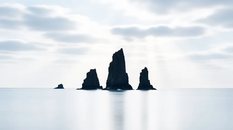 Dark basalt sea stacks emerge from glassy pale water beneath a soft overcast sky.