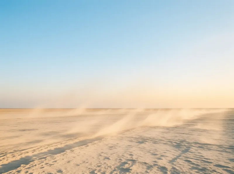 Windblown sand drifts across a vast flat desert floor under a clear blue sky fading to warm golden light near the horizon.