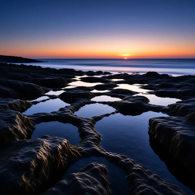 Winding water channels cut through dark coastal rocks while reflecting the soft orange glow of the setting sun.