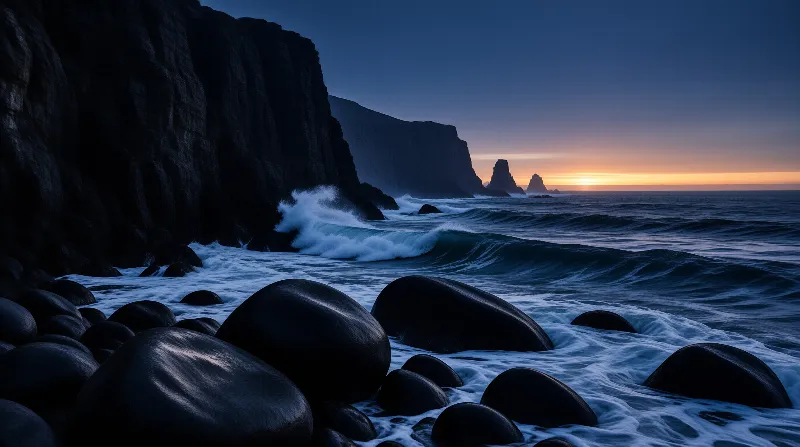 Rounded black boulders rest on a dark sand beach as waves break below towering sea stacks at dusk.