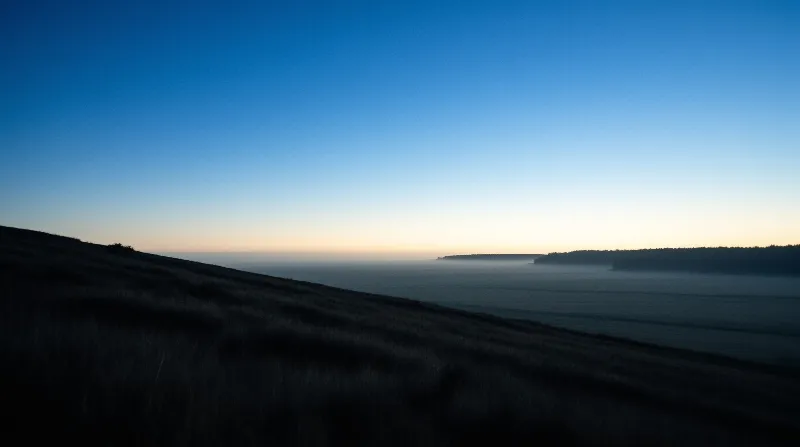 Dark grassy hillside slopes down from the left toward a fog-covered valley beneath a blue to yellow gradient sky at dawn.