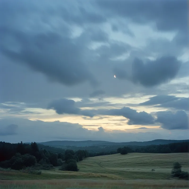 A small crescent moon appears in a cloudy blue sky above rolling grassy fields and distant forested hills during the dim light of twilight.