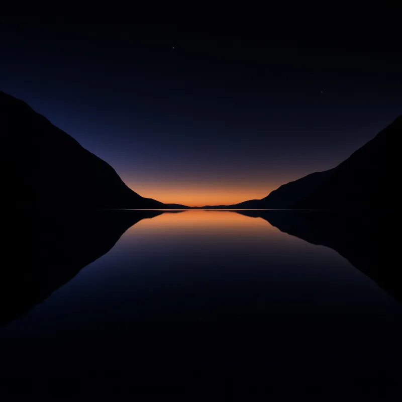 Dark mountain silhouettes frame a calm lake that mirrors the orange glow of the horizon.