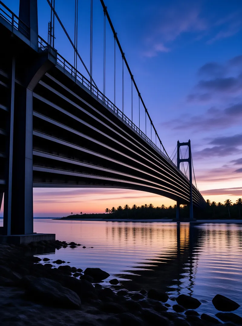 Suspension bridge spanning calm water at dusk with colorful sky reflections and a distant shoreline of palm trees.