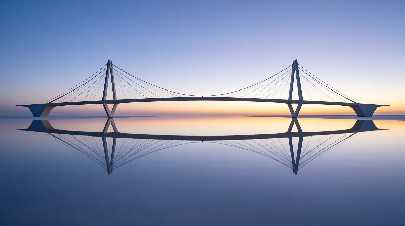 A sleek cable stayed bridge spans calm water at dusk with its towers reflected in a mirror image