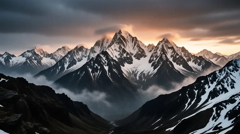 A massive mountain range with sharp snow-covered summits rises above a dark foreground of rocky terrain while low hanging clouds drift through the valleys beneath peaks that catch the warm glow of a setting sun casting long shadows across the rugged landscape.