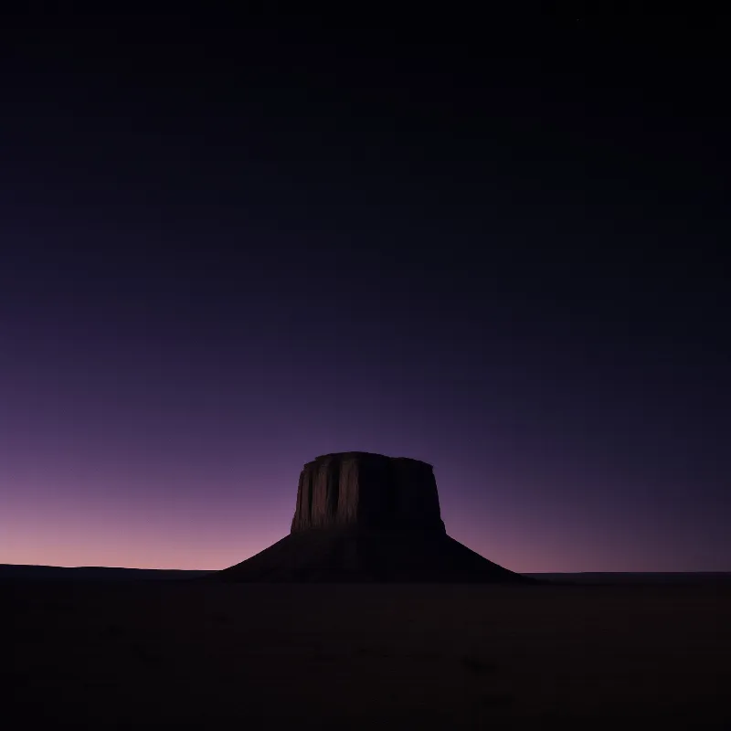 A dark rocky formation stands isolated on the horizon beneath a sky transitioning from deep purple to pink.