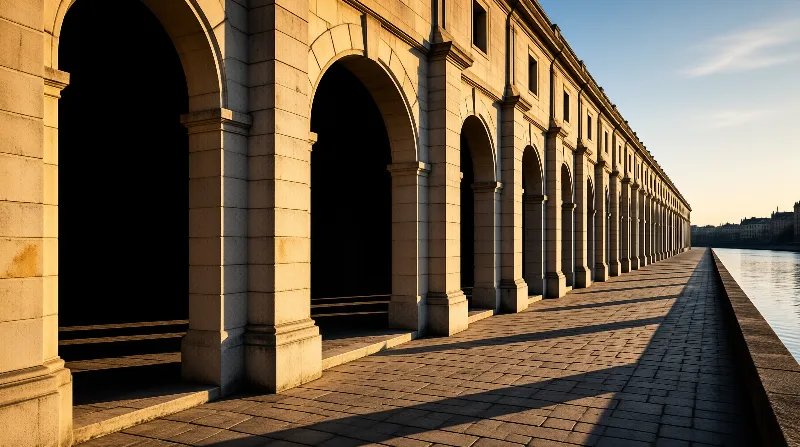 A long series of stone arches lines a paved walkway next to a calm river under warm sunlight.