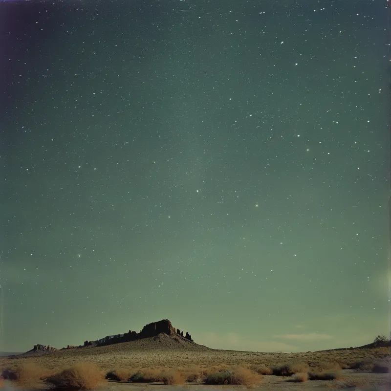 Rocky formation rises from dry desert floor scattered with low shrubs while stars dot a deep teal sky.