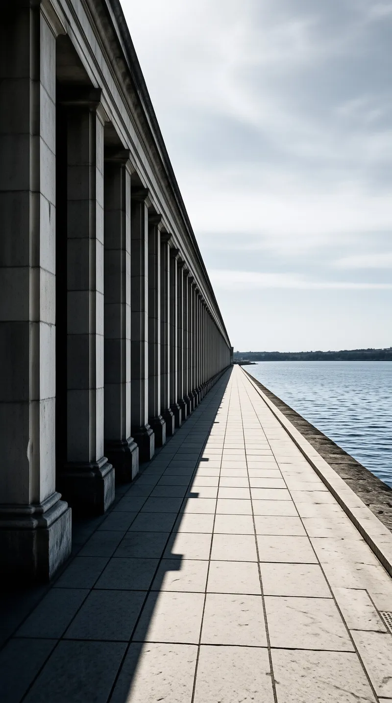 A long arcade of weathered columns shades a tiled promenade that runs beside rippling blue harbor.