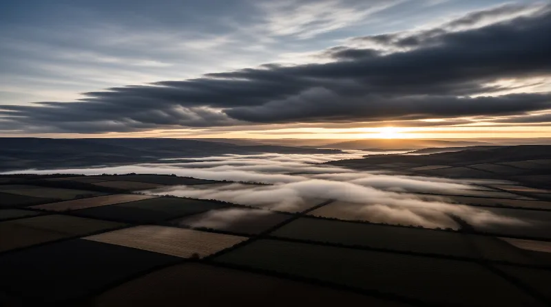 High angle view of agricultural fields with rolling fog banks beneath heavy clouds and a bright sun on the horizon
