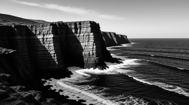 Monochrome coastal cliffs with striped rock layers catch a beam of sunlight above breaking ocean waves