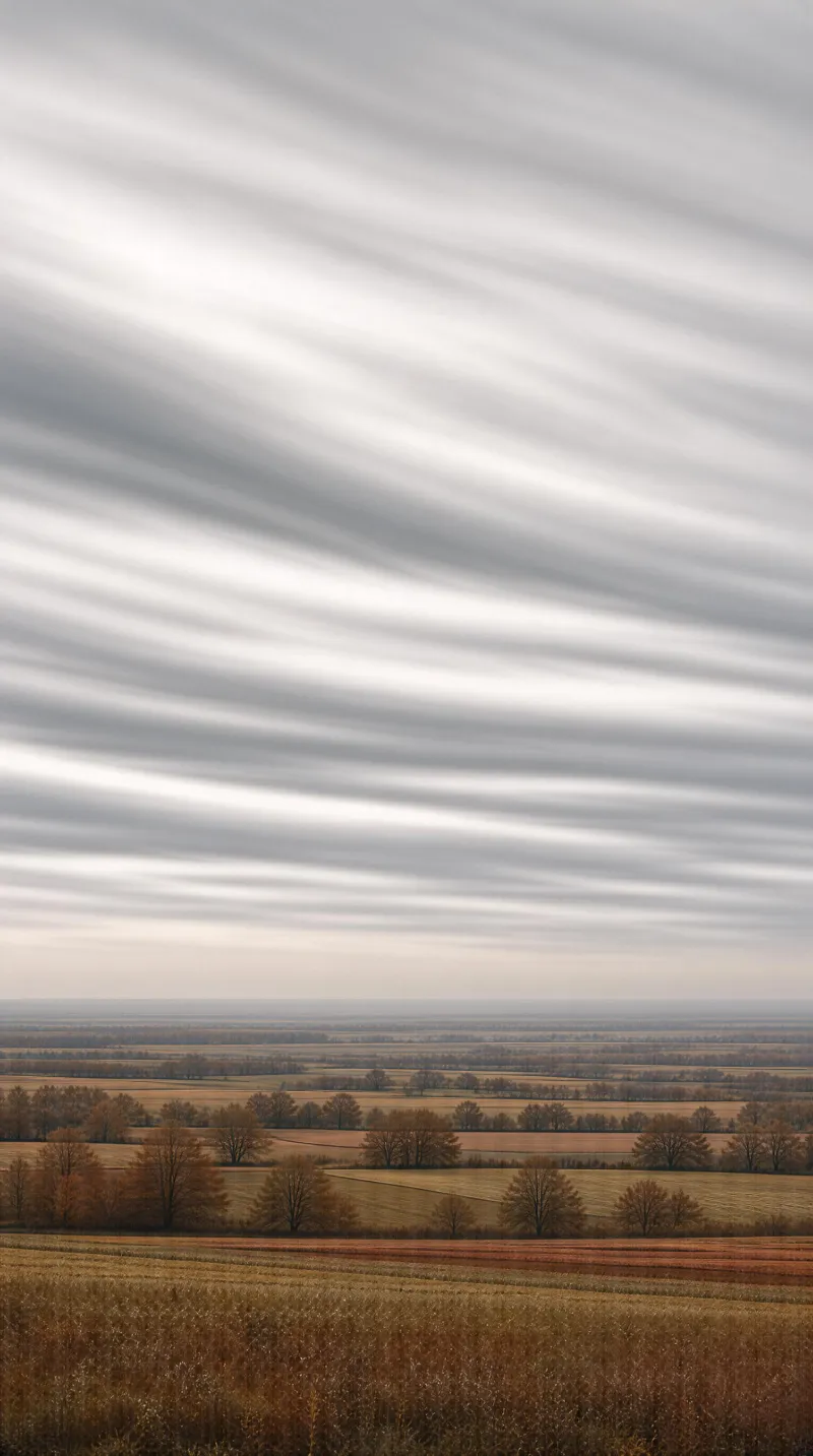 A landscape of open fields with autumn-colored grass and distant trees, set beneath a sky filled with layered, striped clouds.