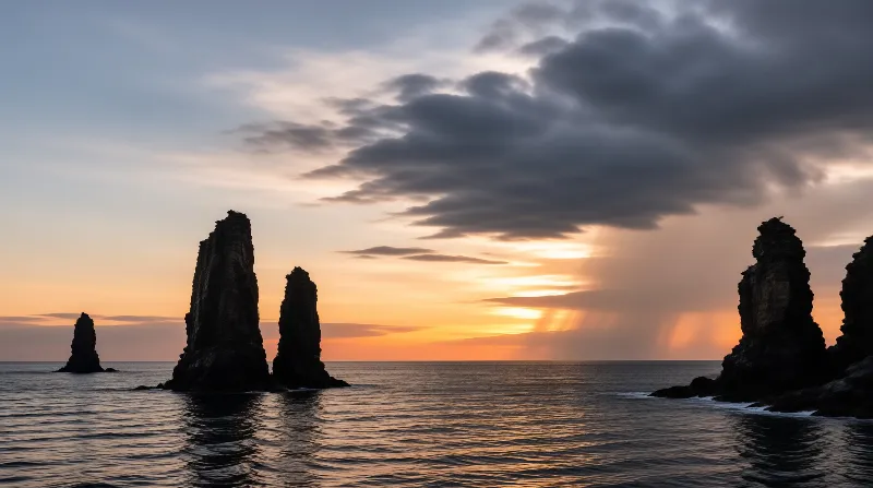 Several dark sea stacks rise from the calm ocean against a vibrant sunset sky with heavy clouds and sunbeams.