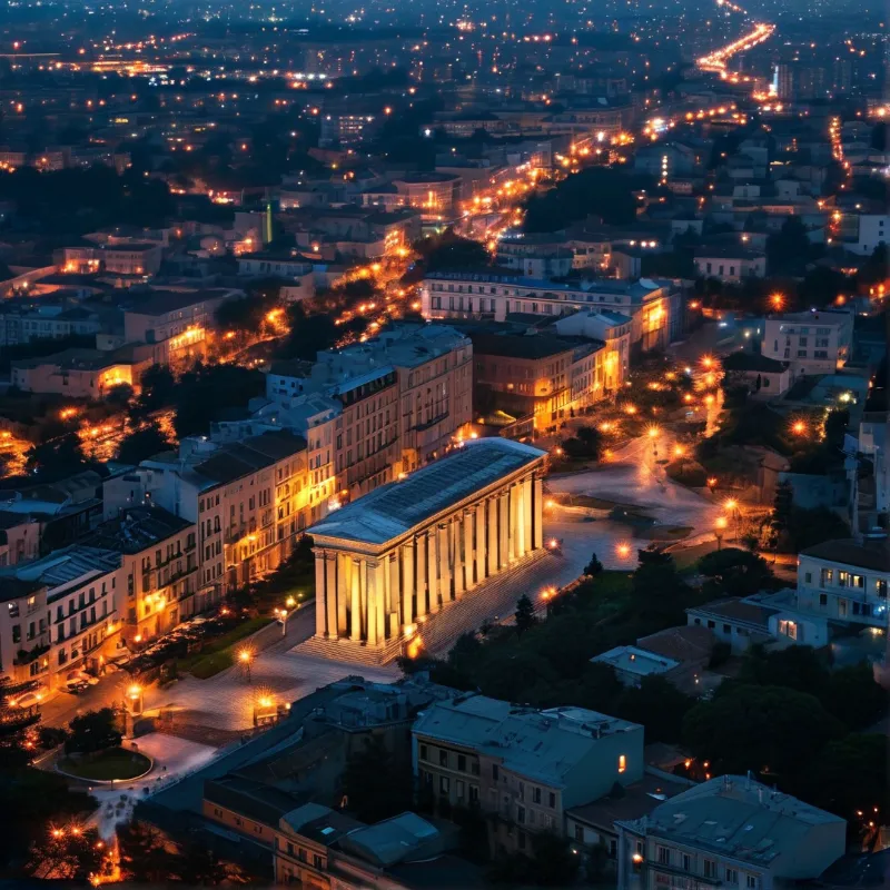 A brightly lit columned structure dominates a sprawling urban landscape filled with warm streetlights and dark trees.