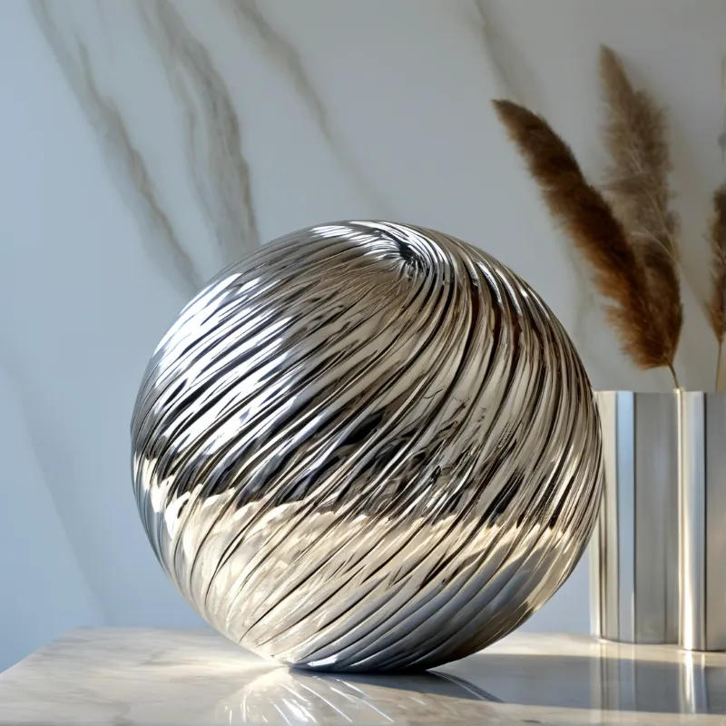 A reflective ridged chrome sphere sits on a polished marble shelf next to a metallic vase with dried grasses under soft light.