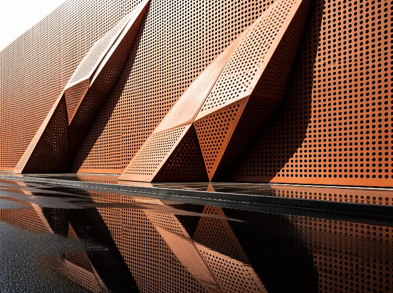 Angled copper panels with a dense pattern of small round holes rise above a dark reflective pool.