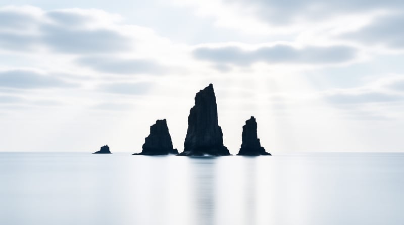 Dark basalt sea stacks emerge from glassy pale water beneath a soft overcast sky.