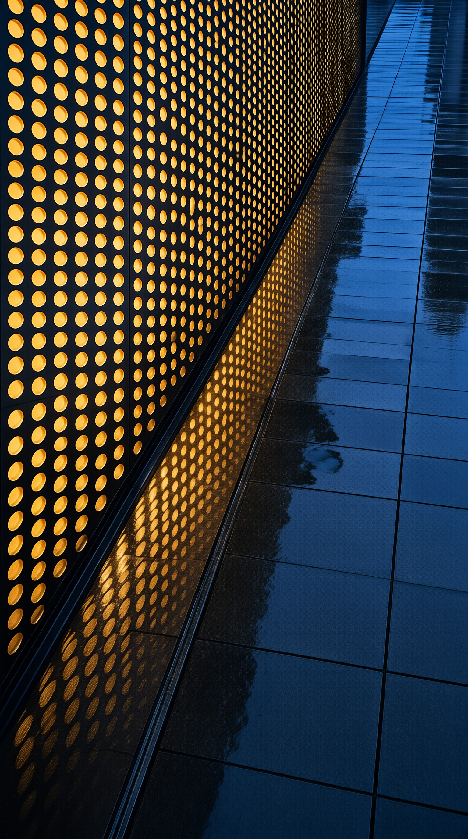 A dark perforated metal panel glows warm orange as its pattern is mirrored on wet deep blue tiled pavement