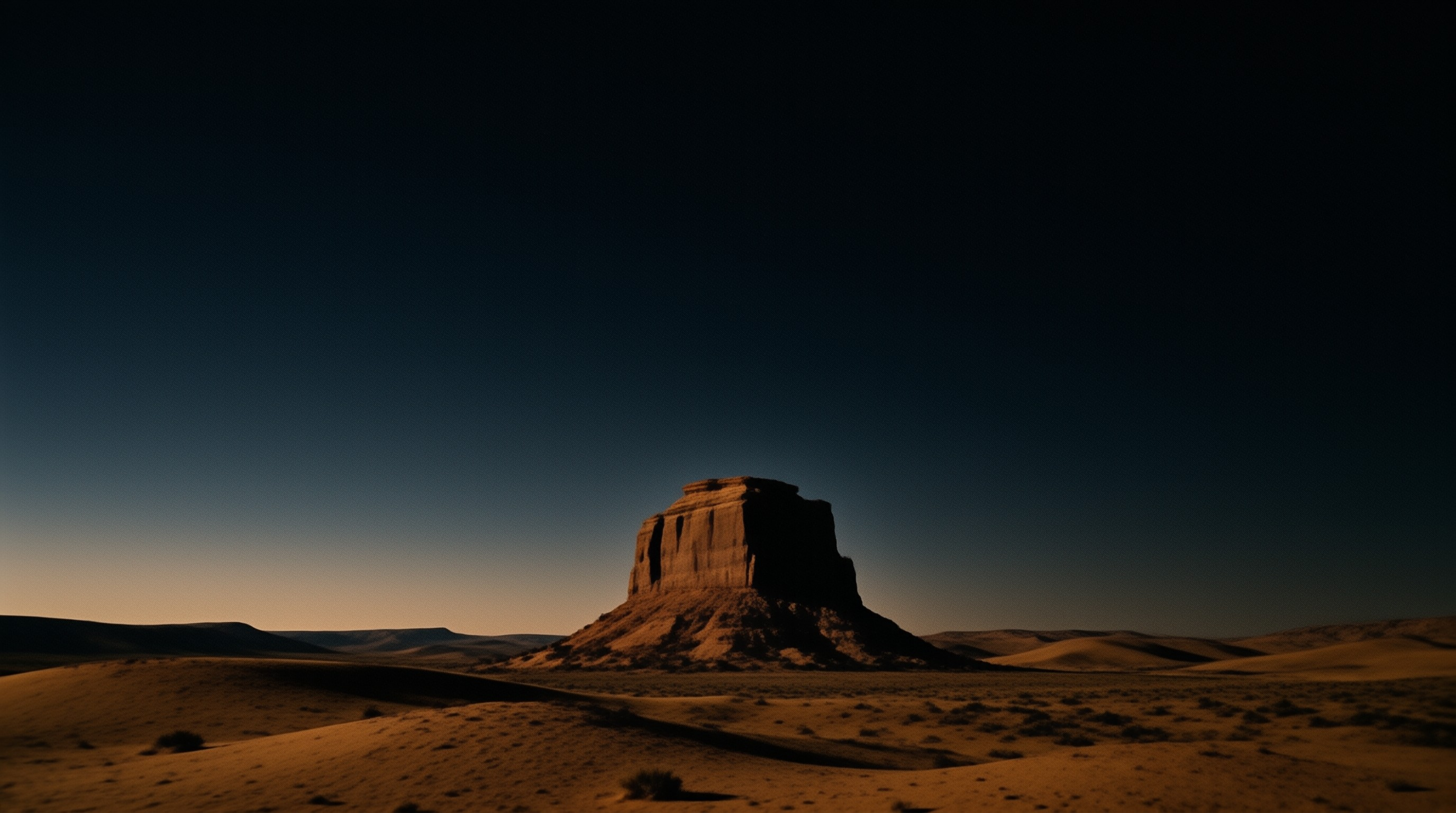 A massive sandstone formation stands in a vast desert landscape, illuminated by warm low sun light against a darkening sky.
