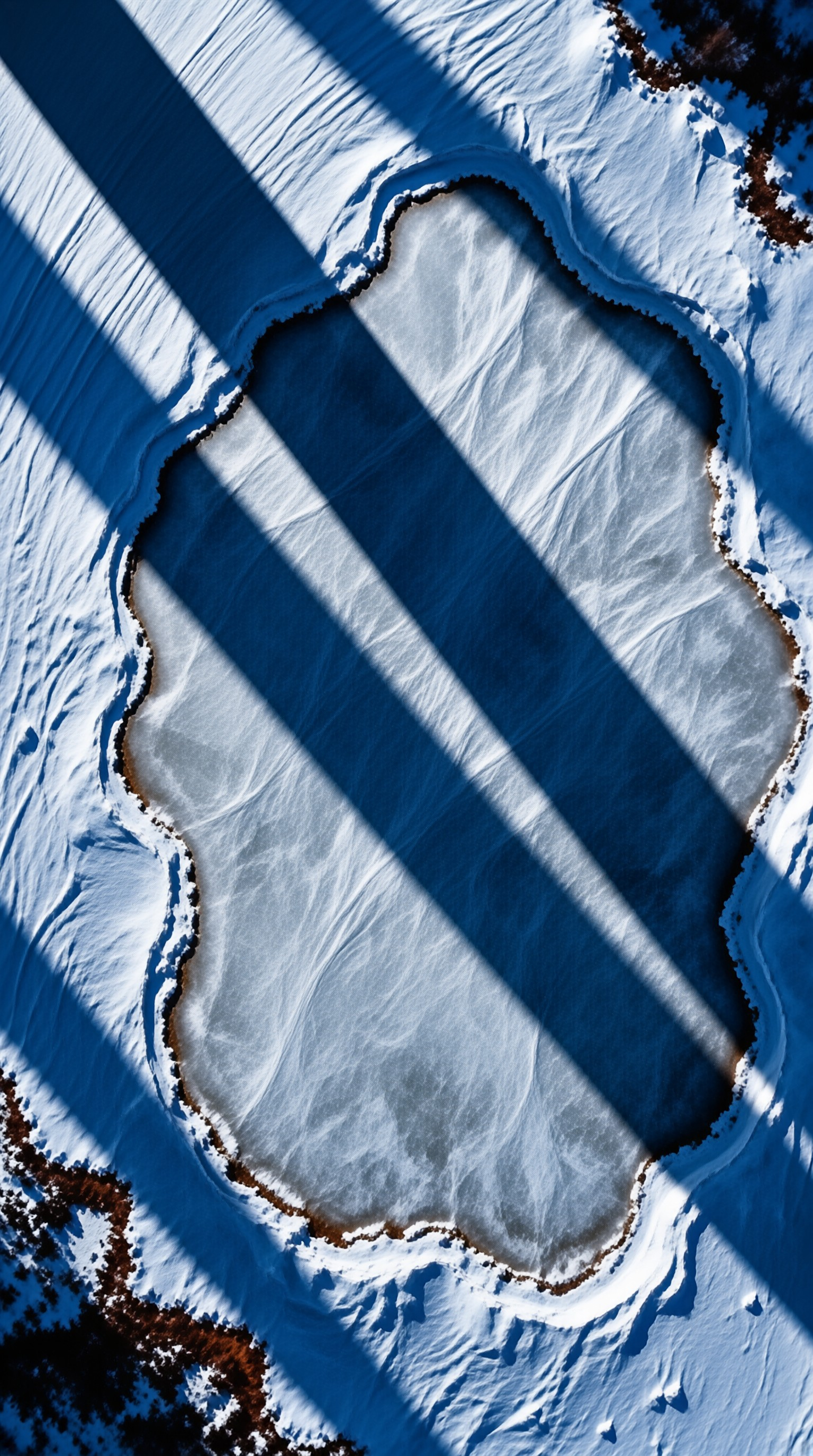 An aerial view of a small frozen lake ringed by snow, crossed by long diagonal shadows cast in cold winter light.