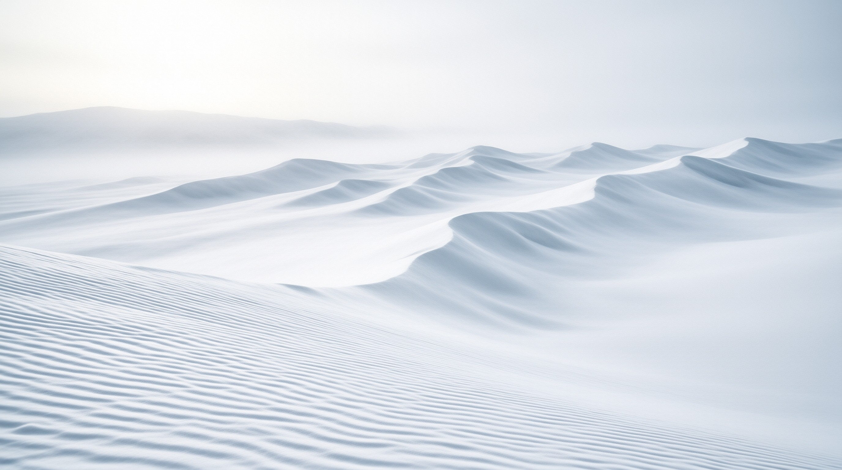 Gentle white sand dunes ripple across the surface while distant ridges disappear into a thick pale fog that softens the entire scene with cool blue tones.