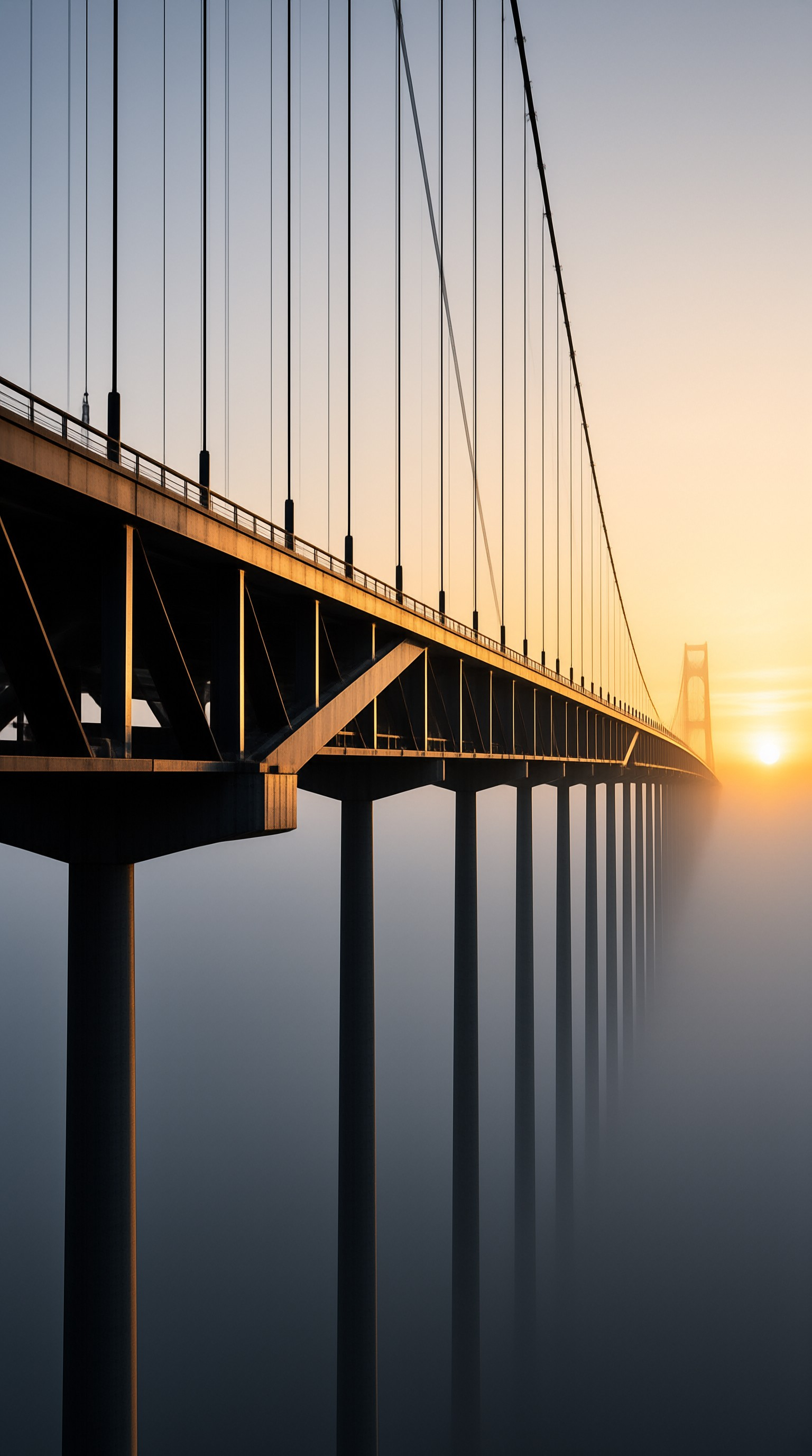 A steel suspension bridge stretches diagonally through thick fog while a bright sun rises on the right horizon.