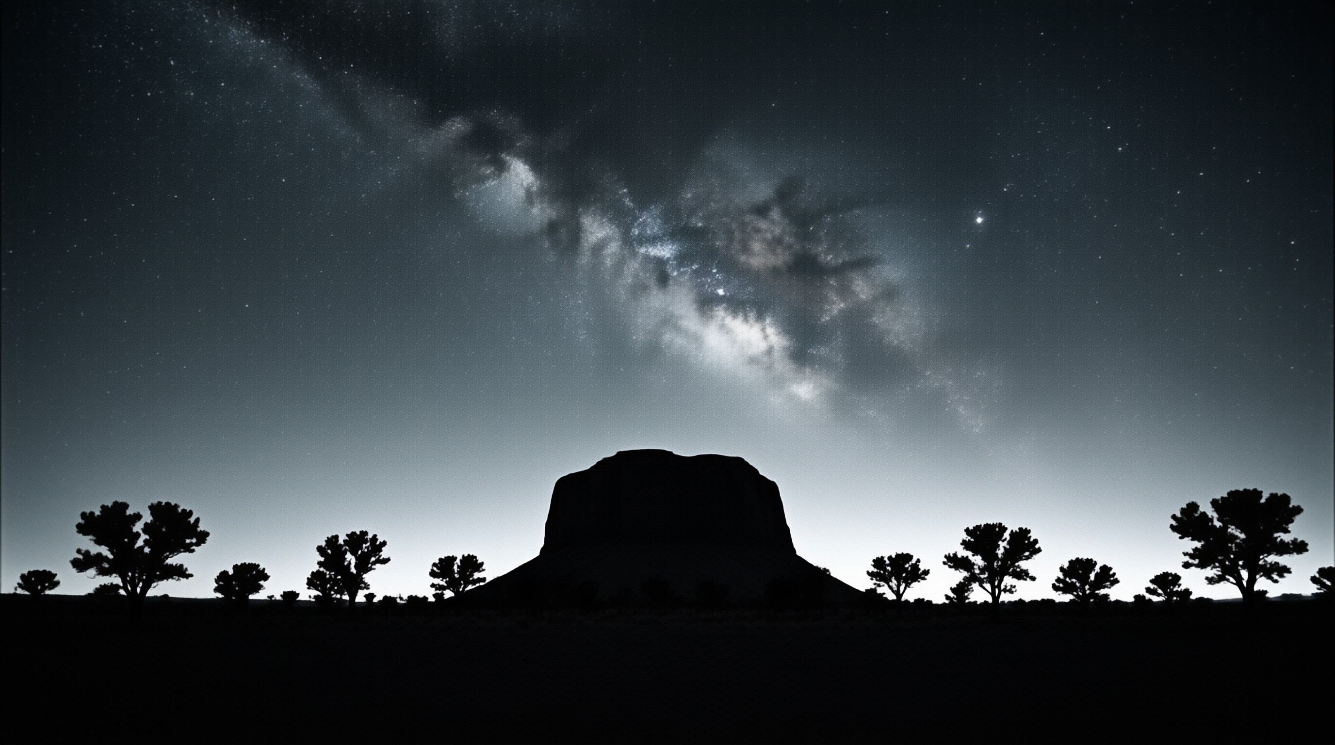 A lone flat-topped mesa and scattered desert trees stand in silhouette under a dense band of stars.